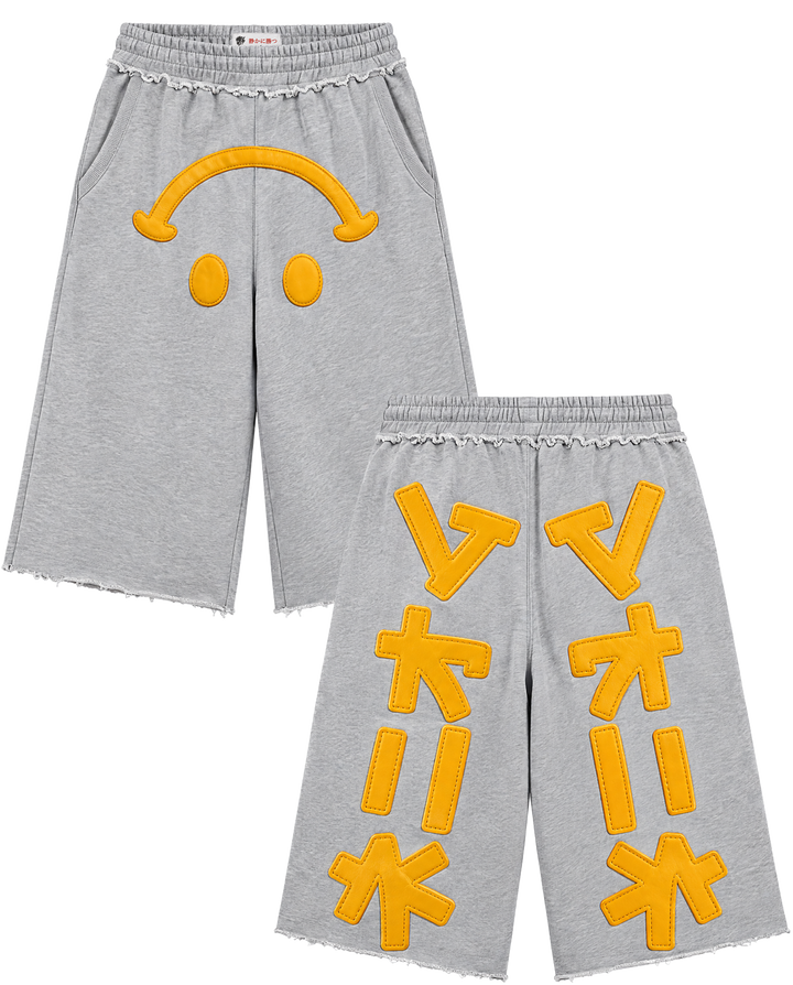 A pair of heather gray cropped sweatpants with yellow applique details. The left pant leg features a yellow smiley face design, and the right pant leg features large yellow geometric letter-like appliqués arranged vertically.