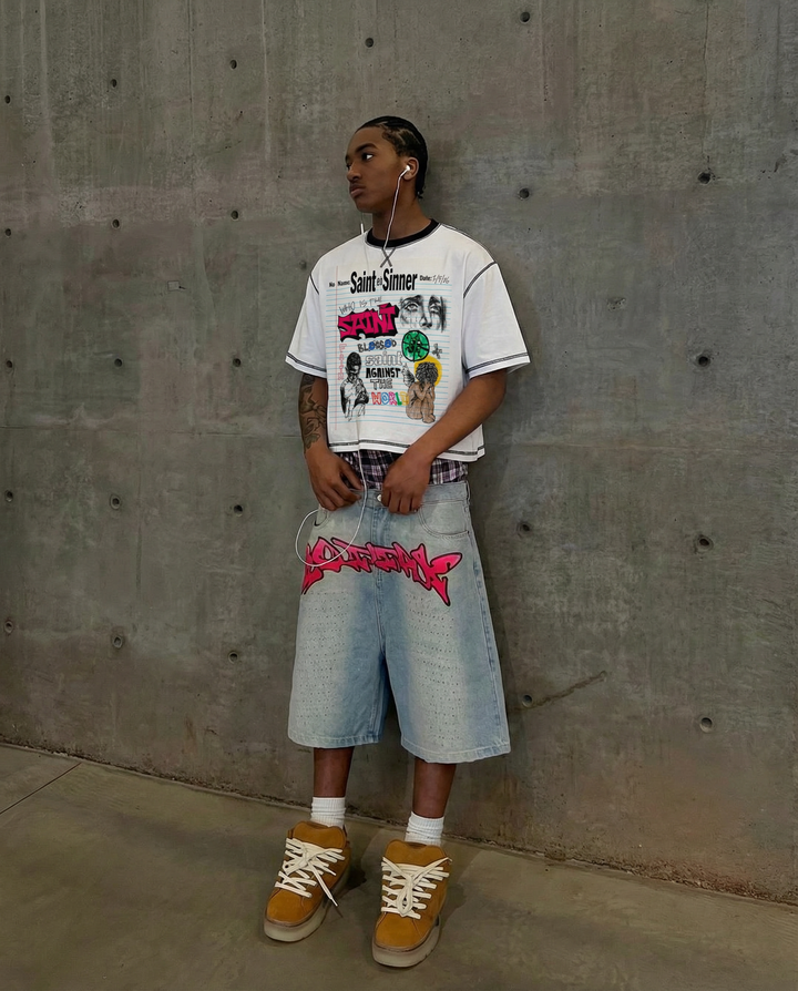 Person wearing a graphic t-shirt and denim shorts against a concrete wall.