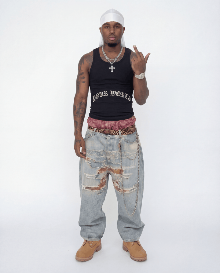 Man wearing a black tank top with text, light blue jeans, and tan shoes on a white background