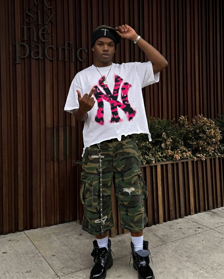 Person wearing a white t-shirt with pink and black design, camouflage shorts, and black boots in front of a wooden wall.