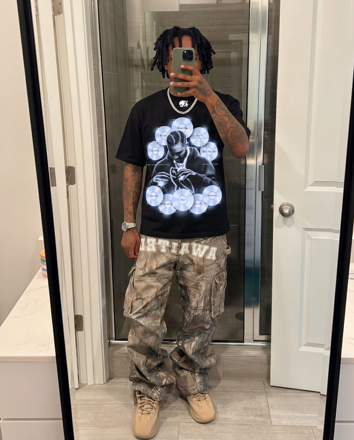 Person wearing a black t-shirt with a graphic design and camouflage pants in a bathroom. Drake Carrer Icon Boxy Shirt Vintage Bootleg Streetwear Oversized Graphic Tee