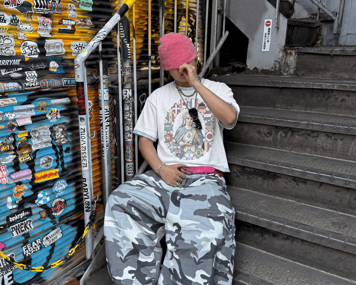 Person sitting on stairs next to a wall with stickers, wearing a white t-shirt and camouflage pants.