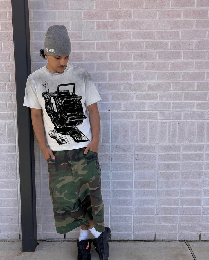 Person wearing a graphic raygun t-shirt, camouflage pants, and a beanie against a brick wall.